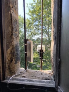 Looking from inside towards two pieces of electrical equipment attached to iron bars outside a window. The left hand equipment is a white box. The right hand equipment is a round glass enclosure on top of a black tube. Both have cables coming from them.