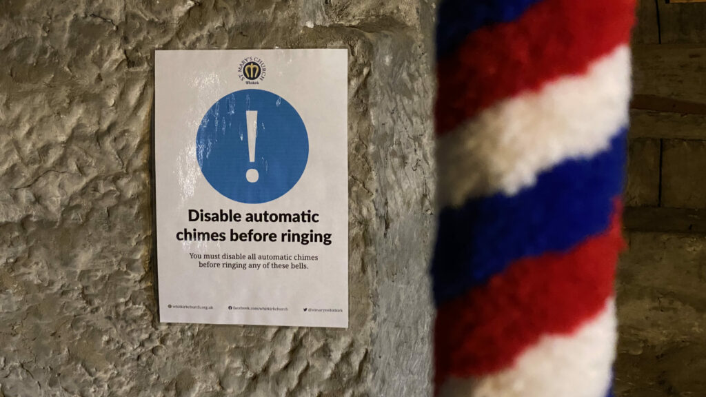 An instruction sign on a stone wall reading "disable automatic chimes before ringing". In the foreground, a red, white and blue stripped portion of a bell rope.