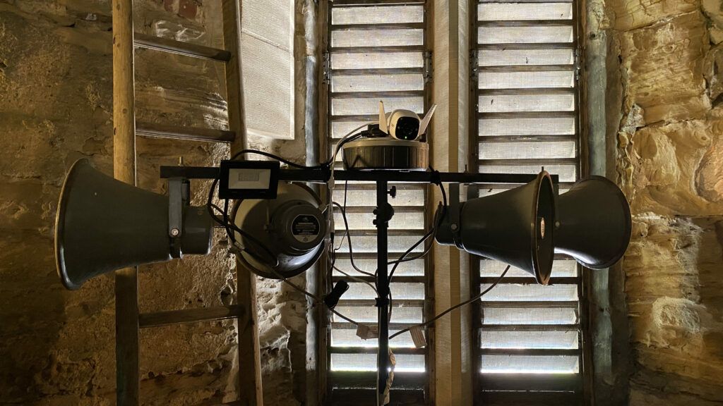 A metal stand with a crossbar on top, holding four speaker horns, a small floodlight, a mains extension and a camera.