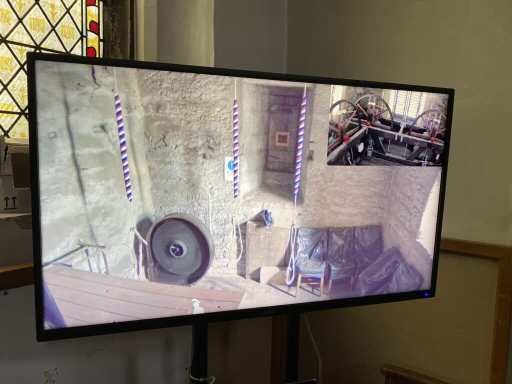 A large television screen showing an image from the ringing room of three bell-ringing ropes. In the top right hand corner there is a picture-in-picture view of the three bells in the belfry.